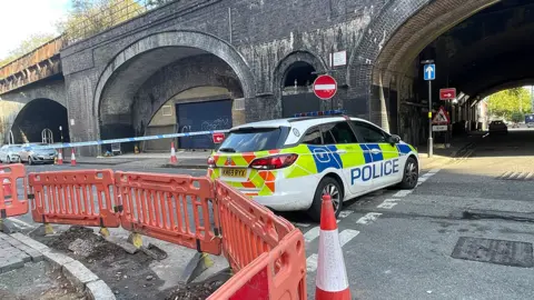 BBC Police car parked across the road, barring it to traffic. Police tape is also across it. The corner also shows orange barriers around roadworks.