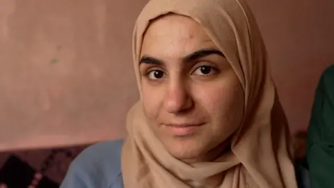 Francesco Tosto/BBC Baraa stares down the barrel of the camera, wearing a beige headscarf, sitting inside a room 