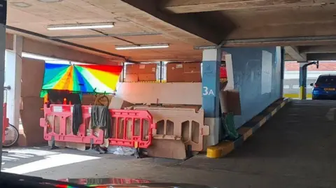 An encampment in a car park in Gloucester. An area of the carpark is bordered off with cardboard and red barriers.