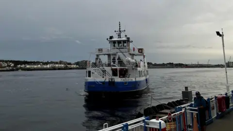 The Shields Ferry, a white and blue boat which has Pride of the Tyne written on the back of it. It has an indoor level, with an outdoor level above it. It is coming into one of the landings which is surrounded by a blue and white fence. One person is waiting by the barriers.
