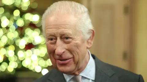 King Charles smiling at an event in the run-up to Christmas 2024, with a lit Christmas tree in the background. 