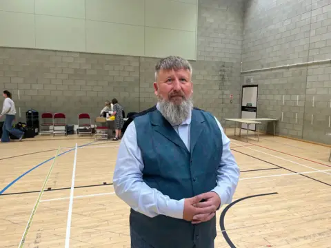 Newly elected Reform councillor Graham Harrison posing for a photo at a leisure centre where the election count has taken place. He is wearing a grey waistcoat and light blue shirt. He has short grey hair and long beard. He is clasping his hands together in front on him and looking at the camera with a neutral expression.