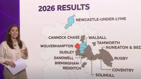 A woman holding a piece of paper is standing next to a map of the West Midlands with several local authority areas highlighted in grey while one is coloured in red and three others are a light shade of blue. All the rest are white with faint grey outlines showing their layout.