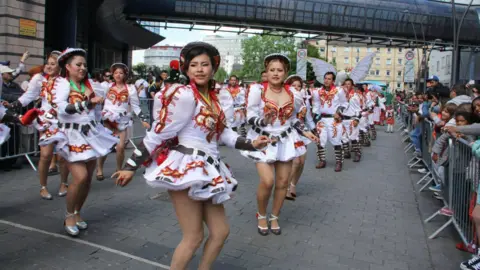 Dozens of men and women dressed in white outfits with puffy sleeves and red and gold trim, dance near the Galaxy in Luton as crowds watch on behind a barrier.