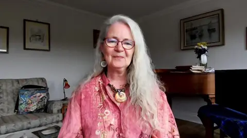 A woman with long white hair and glasses sits facing the camera in a softly lit living room, wearing a pink patterned shirt and a large pendant necklace. Behind her, a wooden desk holds stacked books and a vase of flowers, with framed artwork on the walls and a patterned armchair visible to the left.