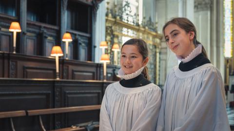 Choristers Lila and Lois pose for press photographers in their surplice inside the cathedral wearing their white ruffed collars and white gowns - known as a surplice - on top of their black cassocks.