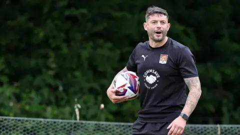 A man with short dark hair and stubble on a football pitch holding a football. He is wearing a black t-shirt, black shorts and a watch. 