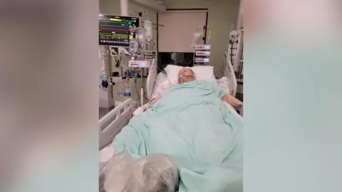 David Taylor A man is lying in an intensive care bed in hospital. He is attached to lots of different medical machines and has an oxygen mask over his face.