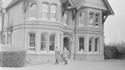 An old black and white photo pf a large two storey building with a triangular roof and big windows. Two men in long coats are walking outside.