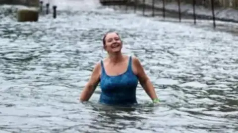 Louise Snelson wearing a blue swimming costume stands in waist-deep water near a slipway. She is looking into the distance and has a big smile on her face. She has a green watch on her left arm.