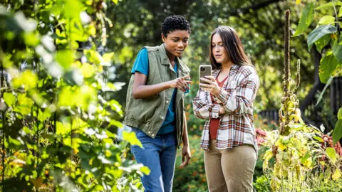 Two young women standing in a garden looking at a mobile phone.