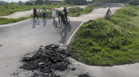 supplied A group of young BMX riders on a track next to a pile of black ash and burnt rubbish.