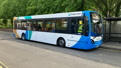 A Stagecoach bus is stopped at a roadside bus stop on a quiet, tree-lined street, with passengers visible inside and a shelter just ahead on the pavement.
