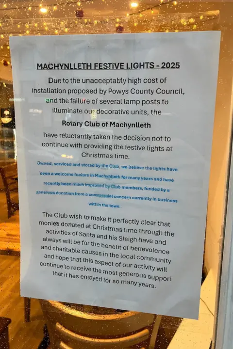 A piece of paper on a shop window. It says 'Machynlleth Festive Lights - 2025' in bold capitals. It continues 'Due to unacceptably high cost of installation proposed by Powys County Council, and the failure of several lamp posts to illuminate our decorative units, the Rotary Club of Machynlleth, have reluctantly taken the decision not to continue providing the festive lights at Christmas time.' It adds that the lights have been a feature feature funded by donations. It also states that money donated through the activities of Santa and his Sleigh have always been donated to local charities. 