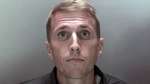 Close up custody photo of Colin Wright - fair haired man with green eyes looking into the camera against a tan coloured background