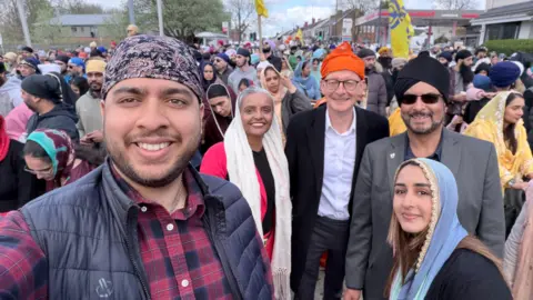 Jules Singh Five people are looking at the camera - it is a selfie, taken by a man in a checked shirt and black gilet and a patterned turban. Next to him is a woman in a bright pink outfit and a cream coloured scarf around her head, the two MPs pictured previously and a woman with a blue scarf and long brown hair. All are smiling.