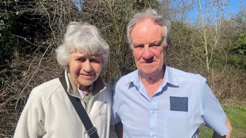 Jane (left) and Peter Middleton - Jane is wearing a light coloured fleece with the strap of a bag over her shoulder. Peter is in a light blue, short-sleeved shirt. behind them are bushes