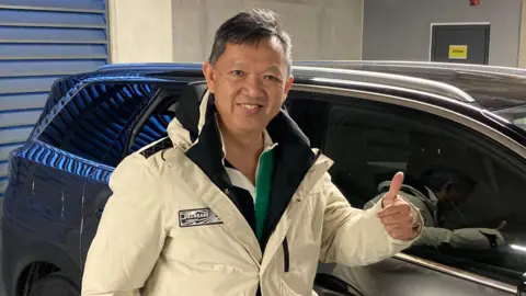 Dave Webster/BBC Ling Yohoya in a cream off white coloured coat, he is looking at the camera and smiling and is giving the camera a thumbs up. He is stood next to a large grey car in a underground car park.