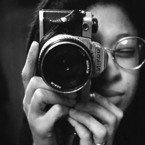 Sandra George Estate A close-up of Sandra, a young black woman with metal-framed glasses, holding a Nikon camera on its side and looking through the view-finder.