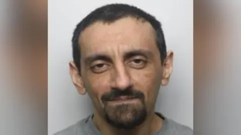 National Crime Agency A mugshot of Hussain. He has short, dark hair and is wearing a light grey crew-neck sweatshirt. The background is plain and light-coloured.