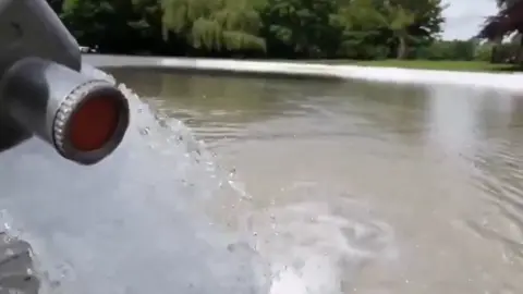 A photo taken from a video showing water going into a pool. Trees and grass are in the background.