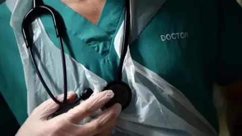 A close up of the chest of a doctor, who is wearing a green vest with the word doctor embroidered onto it. He is wearing white plastic gloves, and a white plastic apron and has a black stethoscope hanging around his neck.