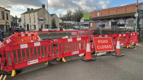 A photo of barriers and a road sign in front of a closed bridge in Lydney in Gloucestershire. Shops and other buildings are visible in the background