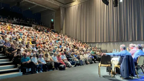 BBC Auditorium style room with hundreds of people sat in a tiered seating area looking at  panel of men in shirts. 