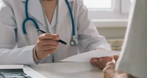 A doctor in a white coat with a blue stethoscope around her neck sits at a desk, photographed from the neck down. They are holding a pen in one hand and a piece of paper in the other.