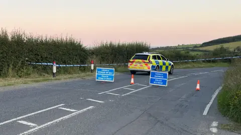 Police car stationary on road which has been cordoned off with blue and white police tape. Blue signs have been put up on the road saying Police Accident and Police Road Closed in English and in Welsh alongside orange and white cones. There is a bend in the road and hills and fields are visible behind.