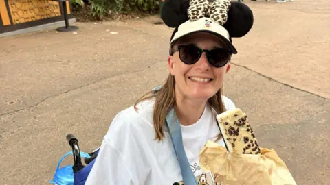 PA Media Jennifer sits in a wheelchair. She is eating a sweet treat from a paper bag and is wearing a white Disney branded T-shirt and a cap with Mickey Mouse ears. She has sunglasses on, her long hair falls below her shoulders, and she is smiling.