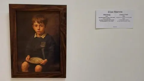 A brown frame that has an image of a school boy holding a round plate on a white wall. There is a white piece of paper next to it that has Elias Marrow and Empty Plate 2024 written on it.