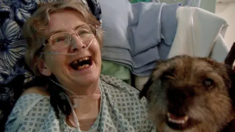 Stephanie, wearing a hospital gown and breathing equipment, has a broad smile on her face as she sits in a chair next to border terrier Hugo, who is looking up at the camera.