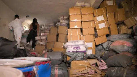 Parks of cardboard boxes and large packages wrapped in plastic stacked to the ceiling inside the warehouse. 