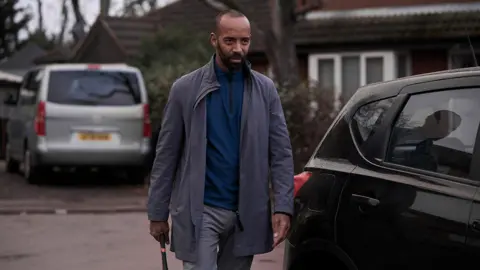 BBC/Left Bank Pictures/James Stack A man with dark hair and a beard is wearing a blue top, grey trousers and a blue jacket as he walks past a black car near a house. A grey van is parked in the background.