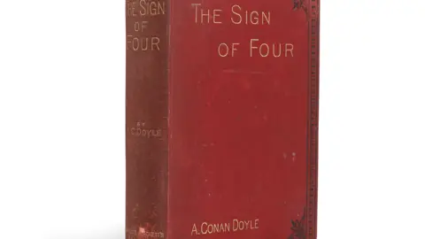 Bonhams A red book with black details on it, reading "The Sign of Four" on both the front and spine, and the author's name "A. Conan Doyle". It is pictured against a plain white background with a shadow behind it.