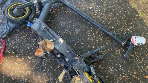 A black e-scooter laid on its side on a road. It has partially crumpled up due to fire damage and has rubbish stuck to parts.