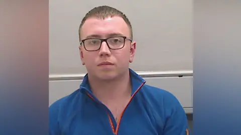 PSNI Cameron Mullen custody image. He has short brown hair wearing glasses and a blue zip up fleece
