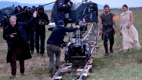 Universal Pictures Christopher Nolan directs at an outdoor shoot for Odysseus - he is standing on the left in black clothing, a camera and crew are in the centre of the image and Matt Damon and Zendaya are on the right