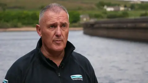A head-and-shoulders image of Dave Kaye. He is a middle-aged man with short grey hair and is wearing a black zip-up fleece featuring the Yorkshire Water logo. Mr Kaye is standing in front of a reservoir.