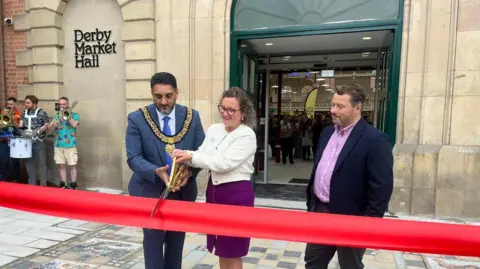 Derby City Council leader cutting a large red ribbon with a pair of large gold scissors outside the entrance of Derby Market Hall, a Grade II listed building the city centre
