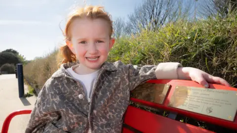 A little girl with red hair, wearing a white t shirt and leopard jacket, sat on a red bench with a gold plaque on it. Behind her is a green bush and a path.