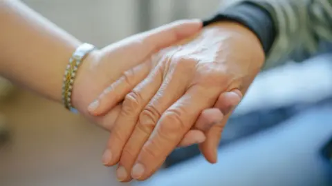 An older person's hand is held by a younger person in a close-up shot. The younger person is wearing a gold and silver bracelet. The older person's forearm is covered by a green and black sleeve. 
