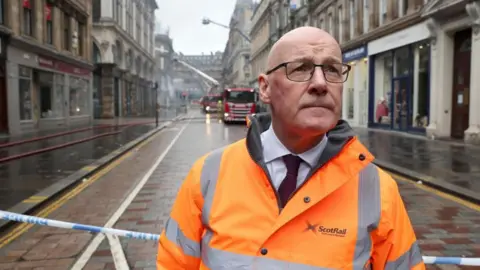 PA Media Swinney wearing a bright orange ScotRail high‑visibility jacket stands in front of an emergency cordon on a wet city street, with fire crews and fire engines working in the background amid smoke