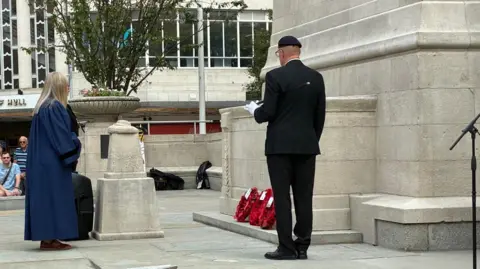 A close up of the stone work which is now a light cream. Two people stand in front of the monument and three red poppy wreaths are at the foot of it.
