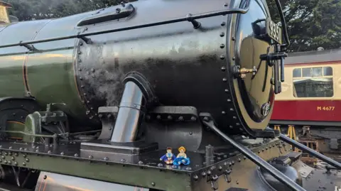 Tabi Payne Two small puppets are sitting on top of a train locomotive.