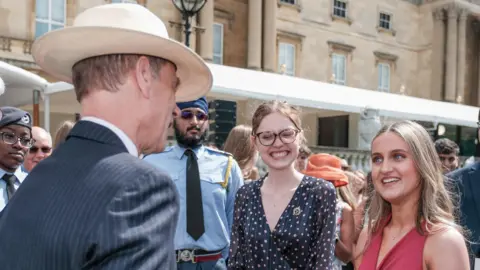 Ian Smither The Duke of Edinburgh, talking to Lucy, at a special garden party, with several people around her. The Duke is wearing a suit pinstriped suit, a hat and has his back to the camera. Zoe is smiling and looking at him.