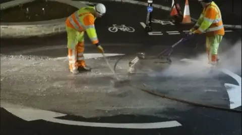 Cathy Hawkins Men in high-vis jackets and trousers use machinery to resurface a roundabout. The roundabout is a hollow circle formed by white arrows