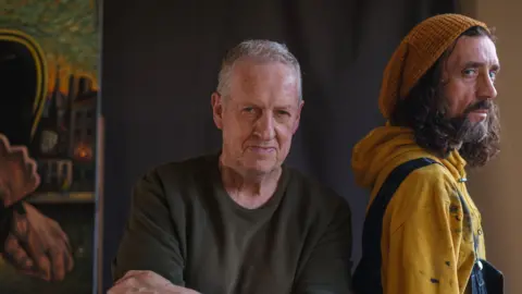 Ross Muir Art Peter Howson, a man in his 60s with short, grey hair is wearing a dark green sweatshirt. He is standing next to Ross Muir, who is in his 40s and has shoulder-length, brown curly hair, and a beard and moustache. He is wearing a yellow beanie hat, a yellow hoodie with splashes of paint on it, and blue denim overalls.