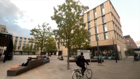 Wide view of an large, open, concreted space in Eddington between buildings with a few trees planted in the central area. There are benches placed randomly, and a man is riding his bike past the area.